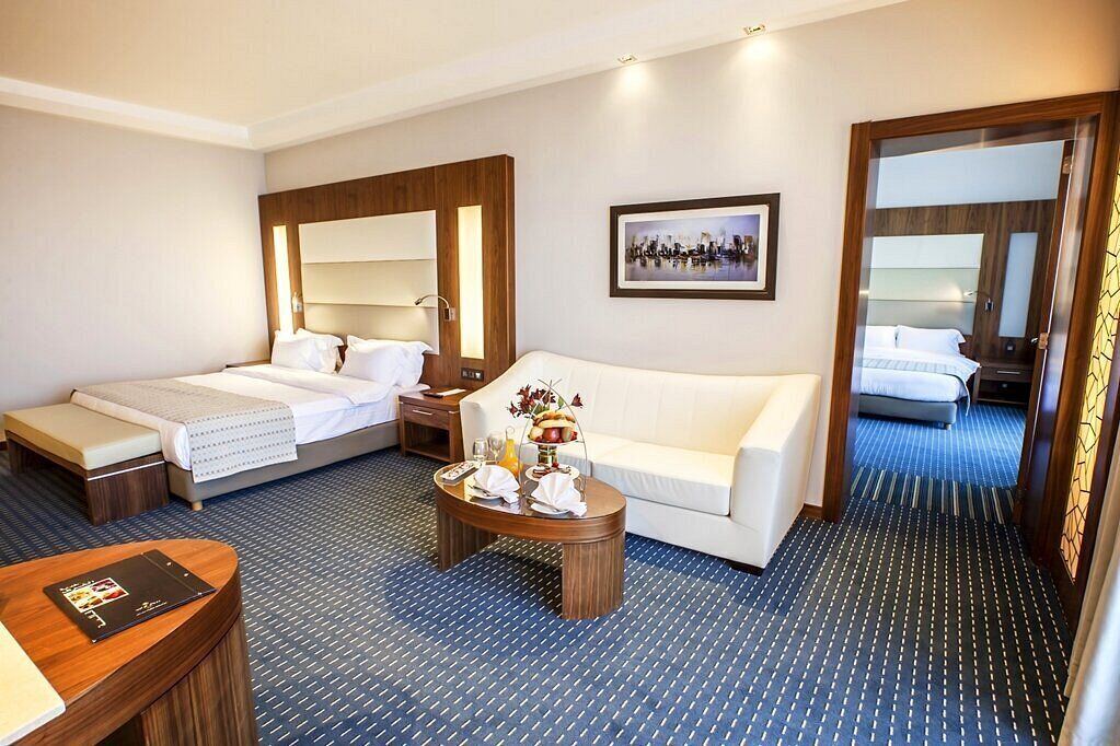 A hotel room with two beds, a white sofa, a small coffee table with breakfast items, a framed picture on the wall, and a patterned carpet.