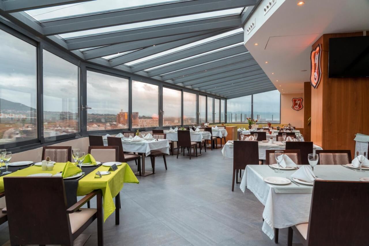 Un restaurant où les tables sont dressées pour le service, de grandes fenêtres offrant une vue sur la ville et un décor intérieur moderne.