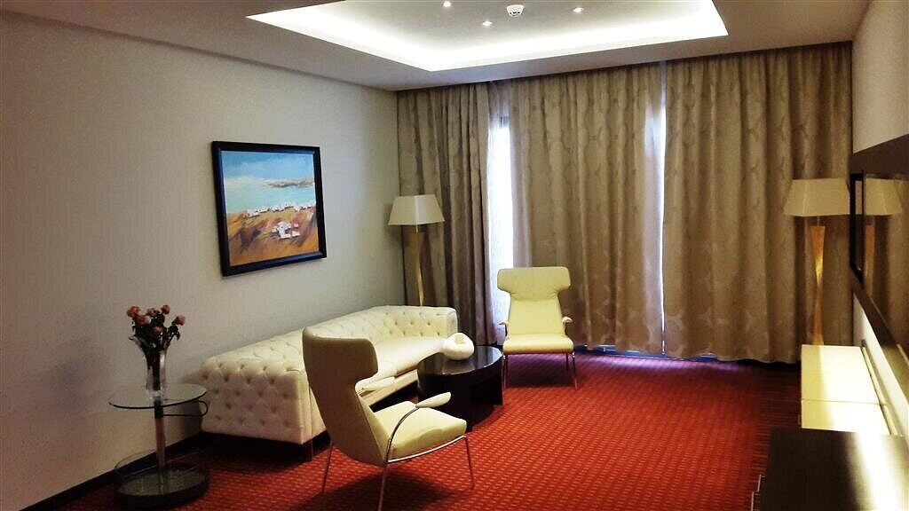 Habitación de hotel con alfombra roja, un sofá blanco, un sillón amarillo, una mesita redonda y un cuadro de una playa.