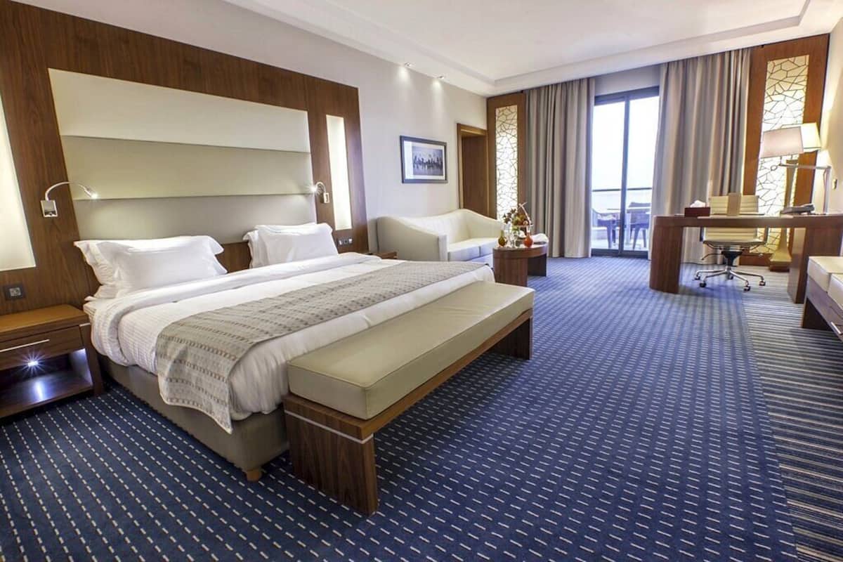 Habitación de hotel con una cama grande, un escritorio con silla, un pequeño sofá y vistas al exterior a través de un ventanal.