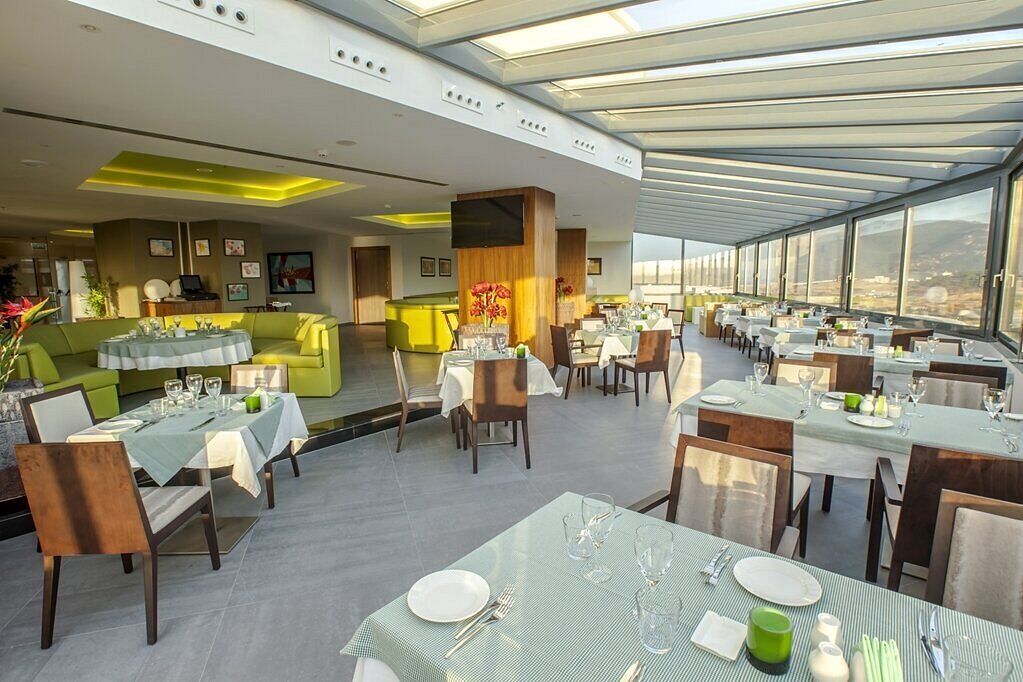 Un restaurante moderno con techo alto y ventanas amplias, que presenta una mezcla de mesas de comedor y un área de estar con un sofá verde.