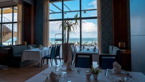 A restaurant with a view of the ocean, featuring a table set for dining, chairs, and a large potted plant. - Royal Tulip Skikda (Filfila)