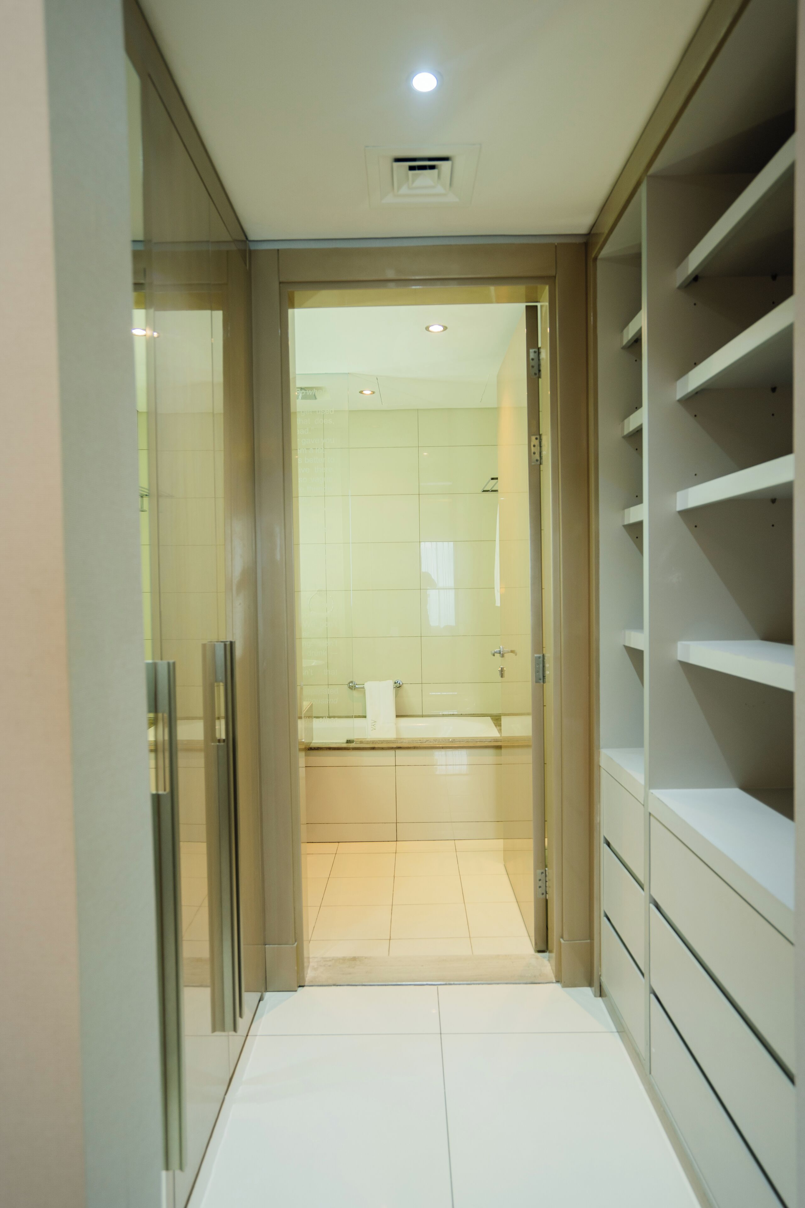A narrow hallway leading to a bathroom with a toilet, sink, and towel rack. There are shelves on the right side and a door on the left.