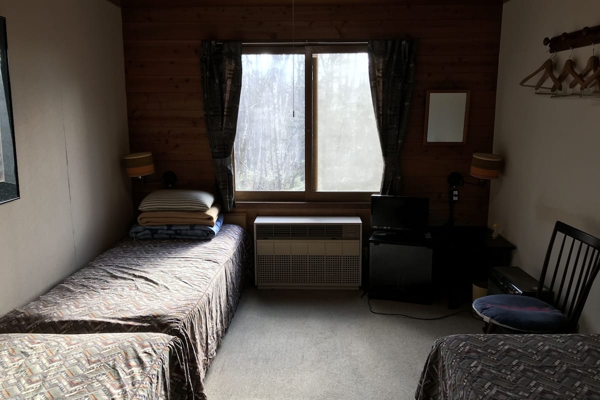 A small, cozy room with two beds, a desk, and a chair. There is a window with curtains, a heater, and a mirror on the wall.