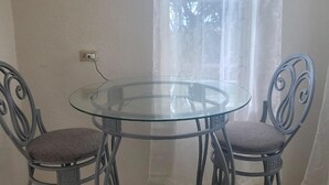 Ein kleiner Glastisch mit zwei Metallstühlen, eine Steckdose an der Wand und ein Fenster mit durchscheinenden Vorhängen.