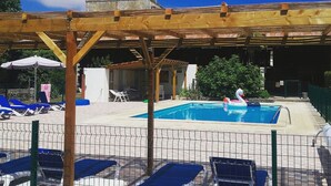 Uima-allasalue, jossa on puinen pergola, aurinkotuoleja ja kirkkaan sininen taivas.