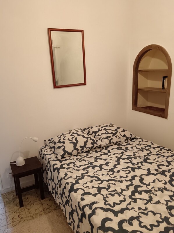 A bedroom with a bed, a bedside table with a lamp, a mirror, and a small window.