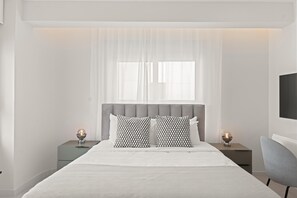 Une chambre moderne avec un grand lit, deux tables de chevet avec des lampes, une tête de lit rembourrée de couleur grise et une grande fenêtre ornée de rideaux légers.