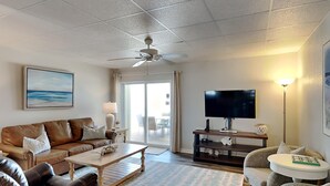 Una sala de estar con un sofá de cuero marrón, una mesa de centro de madera, un televisor de pantalla plana sobre un mueble, un ventilador de techo y un ventanal con vistas al exterior.