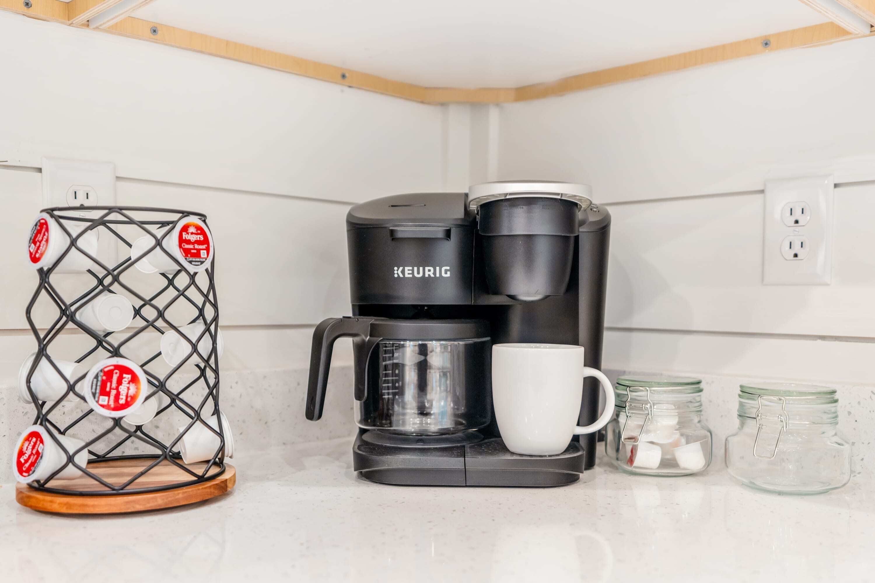 A black Keurig coffee maker with a clear coffee pot and a white mug, a metal basket with coffee pods, and two glass jars with sugar cubes.