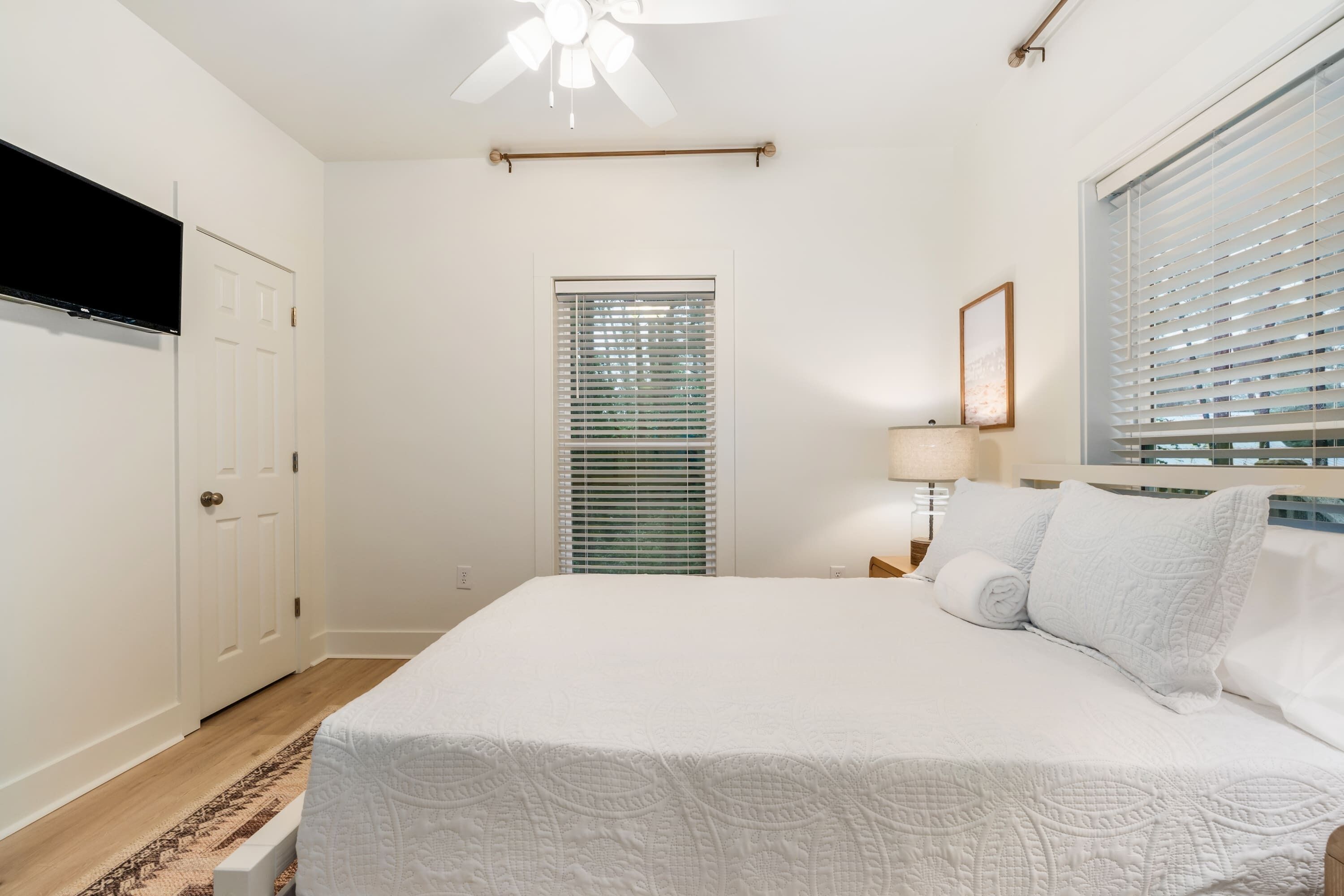 A bedroom with a bed, a ceiling fan, a window with blinds, a door, and a wall-mounted TV.