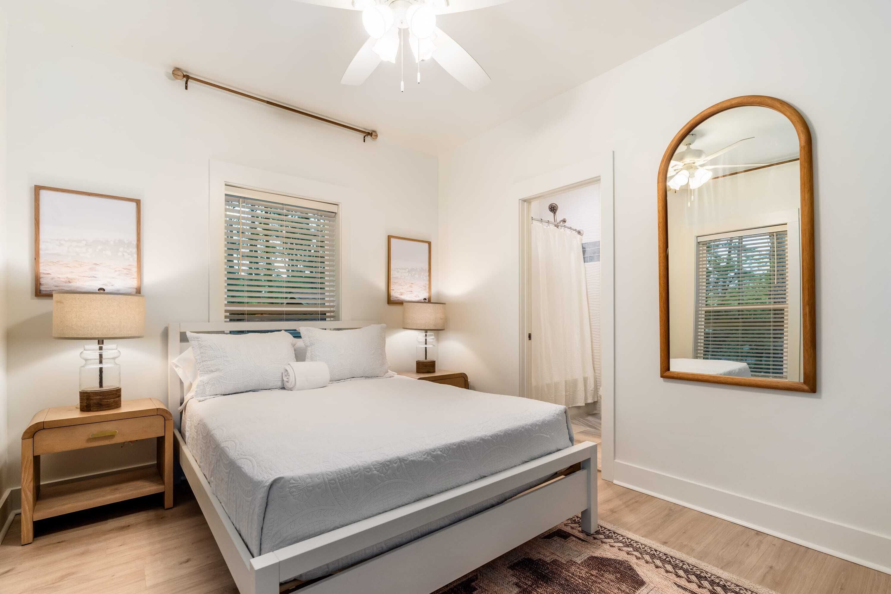 A bedroom with a bed, two bedside tables, a ceiling fan, a large mirror, and a window with blinds.