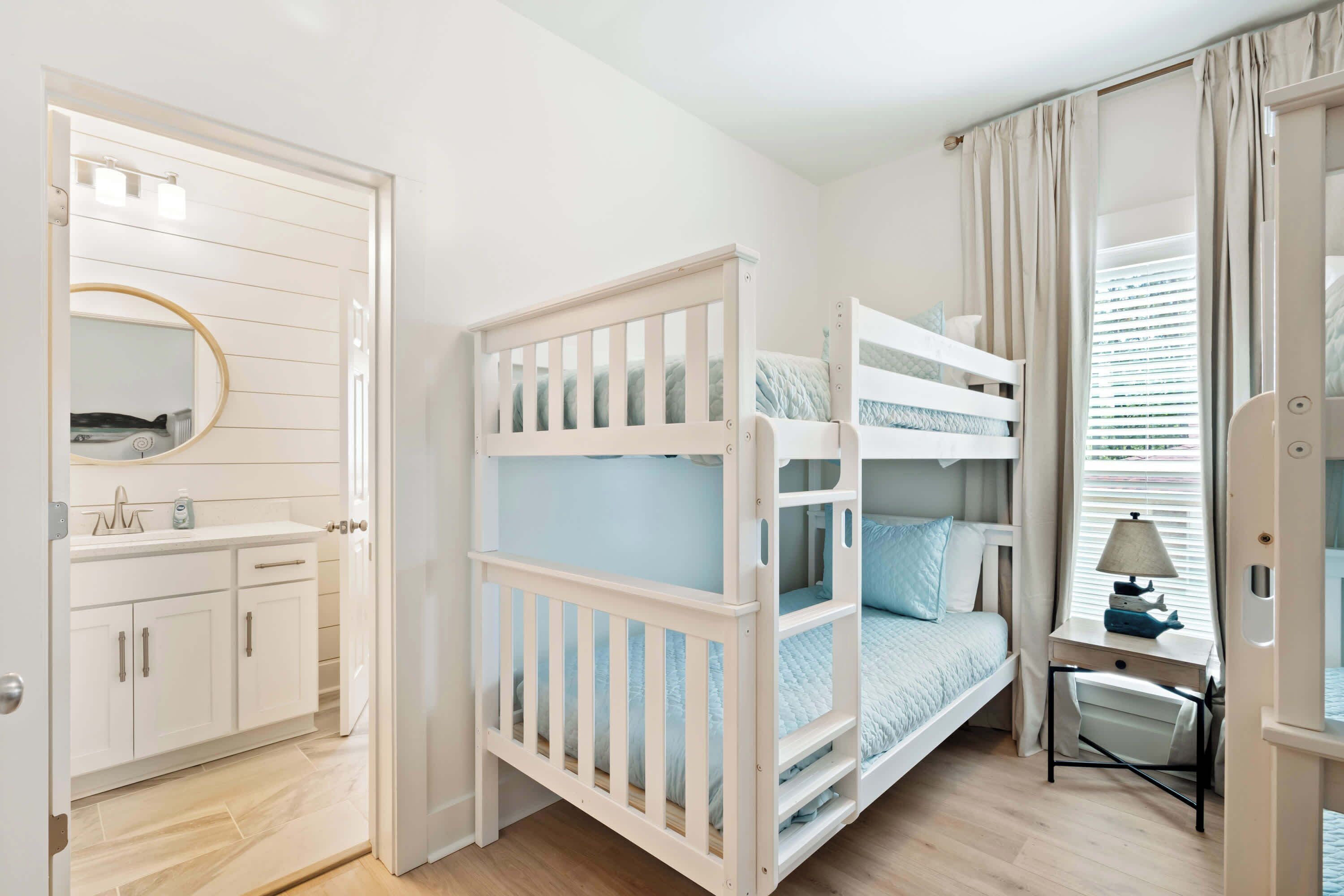 A bedroom with a bunk bed, a bedside table with a lamp, a window with curtains, and a bathroom with a mirror and cabinets.