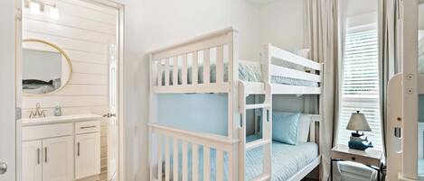 A bedroom with a bunk bed, a bedside table with a lamp, a window with curtains, and a bathroom with a mirror and cabinets.