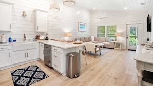 Une cuisine moderne avec des meubles blancs, un îlot central, des luminaires suspendus et une vue sur l’extérieur grâce à de grandes fenêtres.
