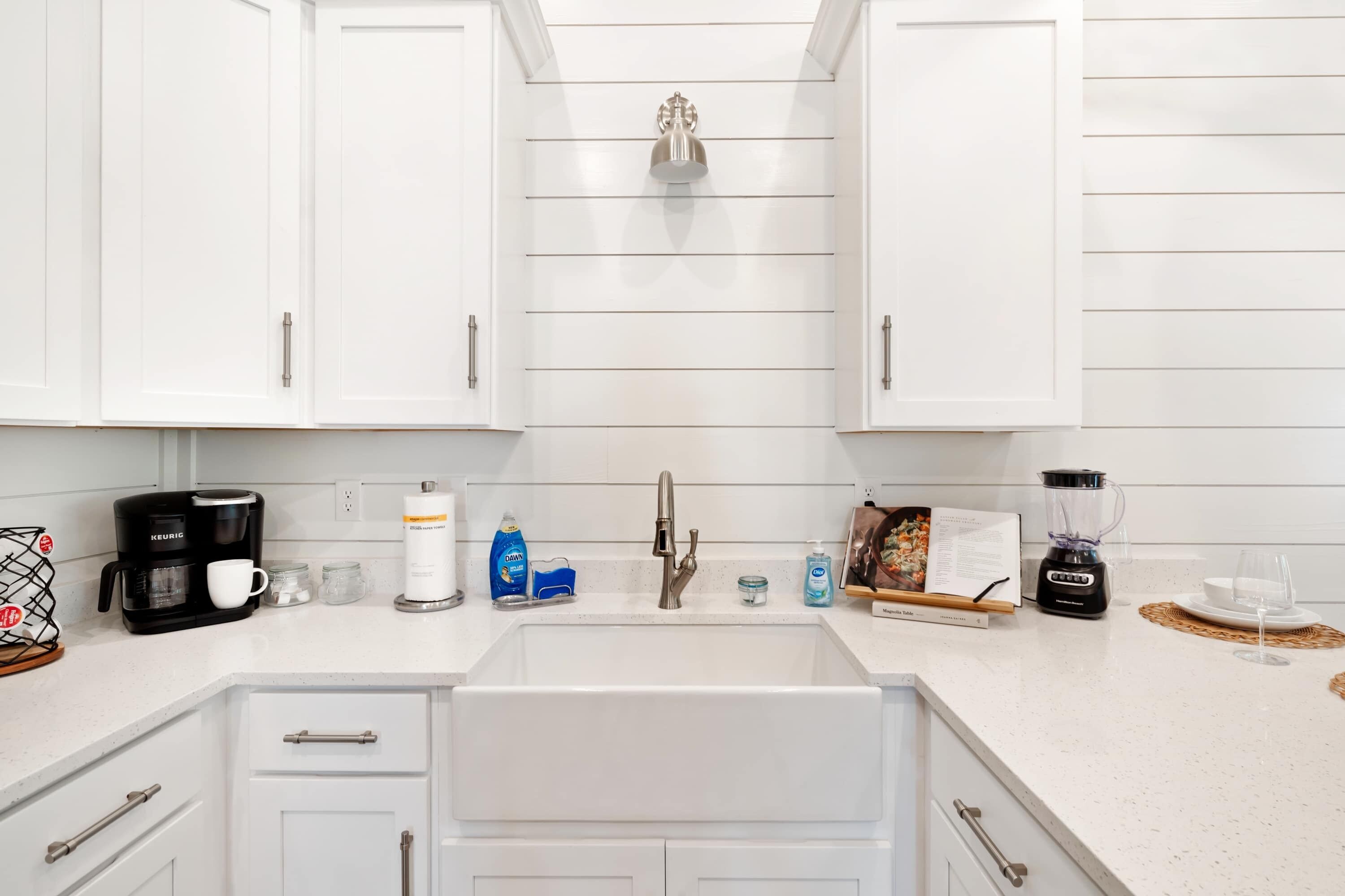 A modern kitchen with white cabinetry, a stainless steel sink, and various appliances including a coffee maker, a blender, and a kettle.
