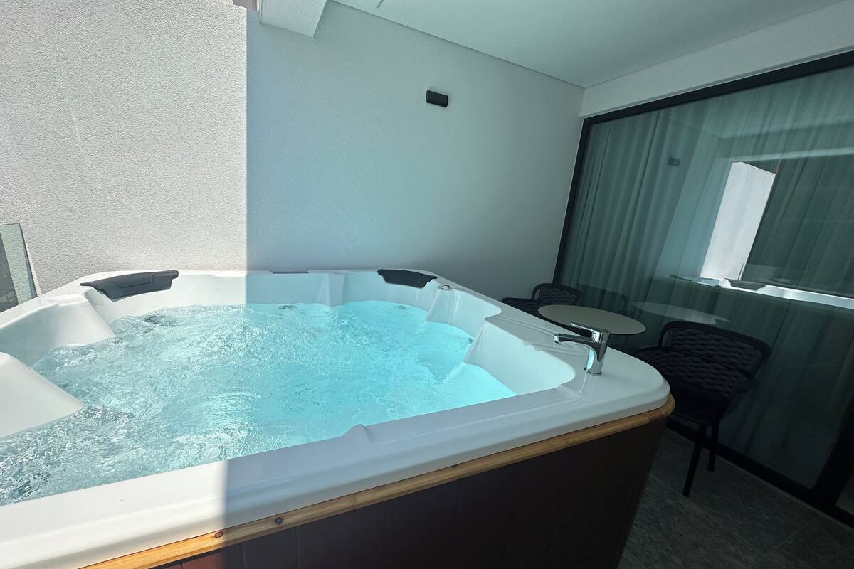 Ein Whirlpool mit Holzrahmen, einem kleinen Tisch und einem Sessel in einem Raum mit Glaswand und Fenster.
