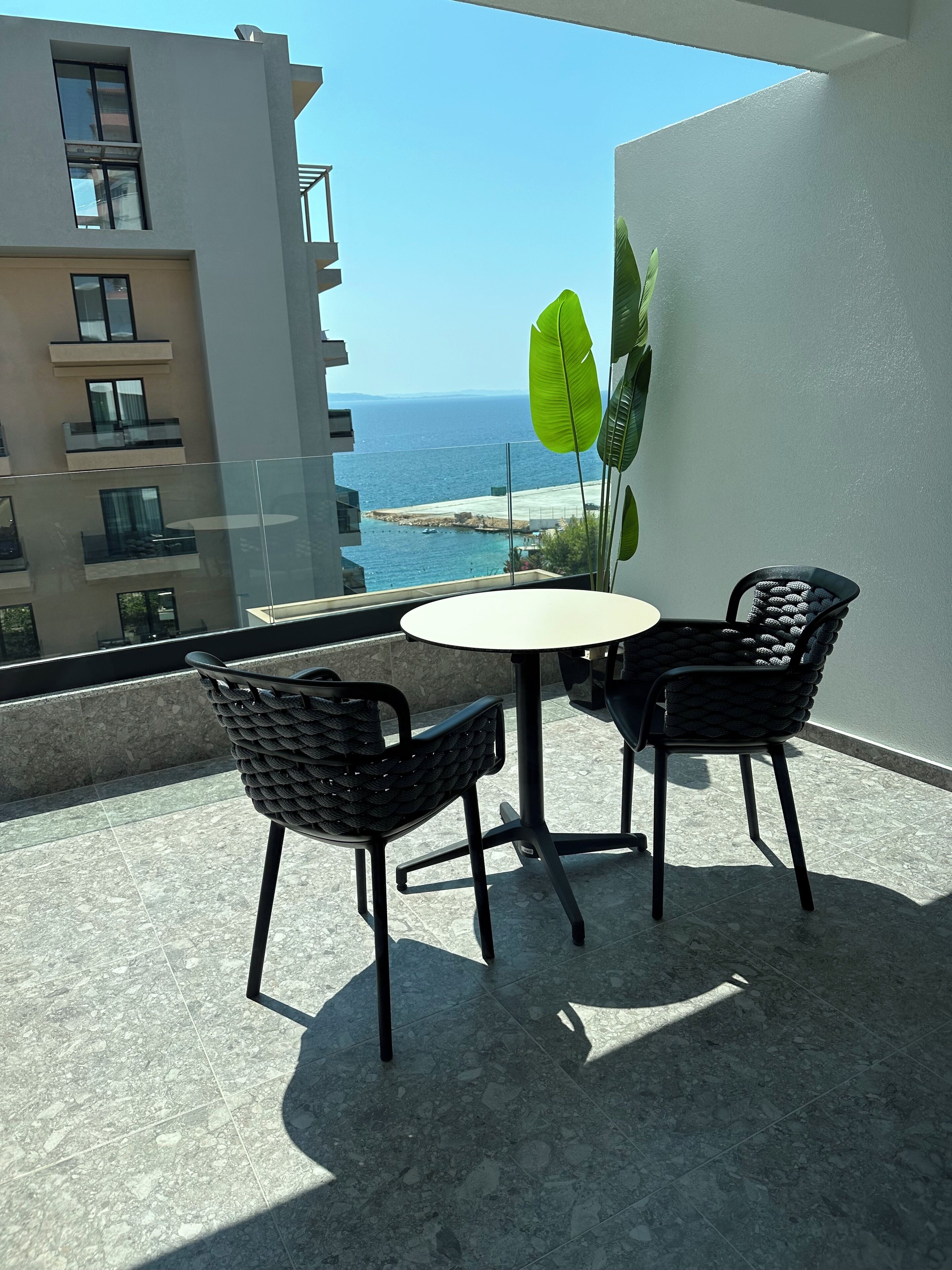 A balcony with a round table and two chairs, overlooking a coastal view with buildings and a clear sky.