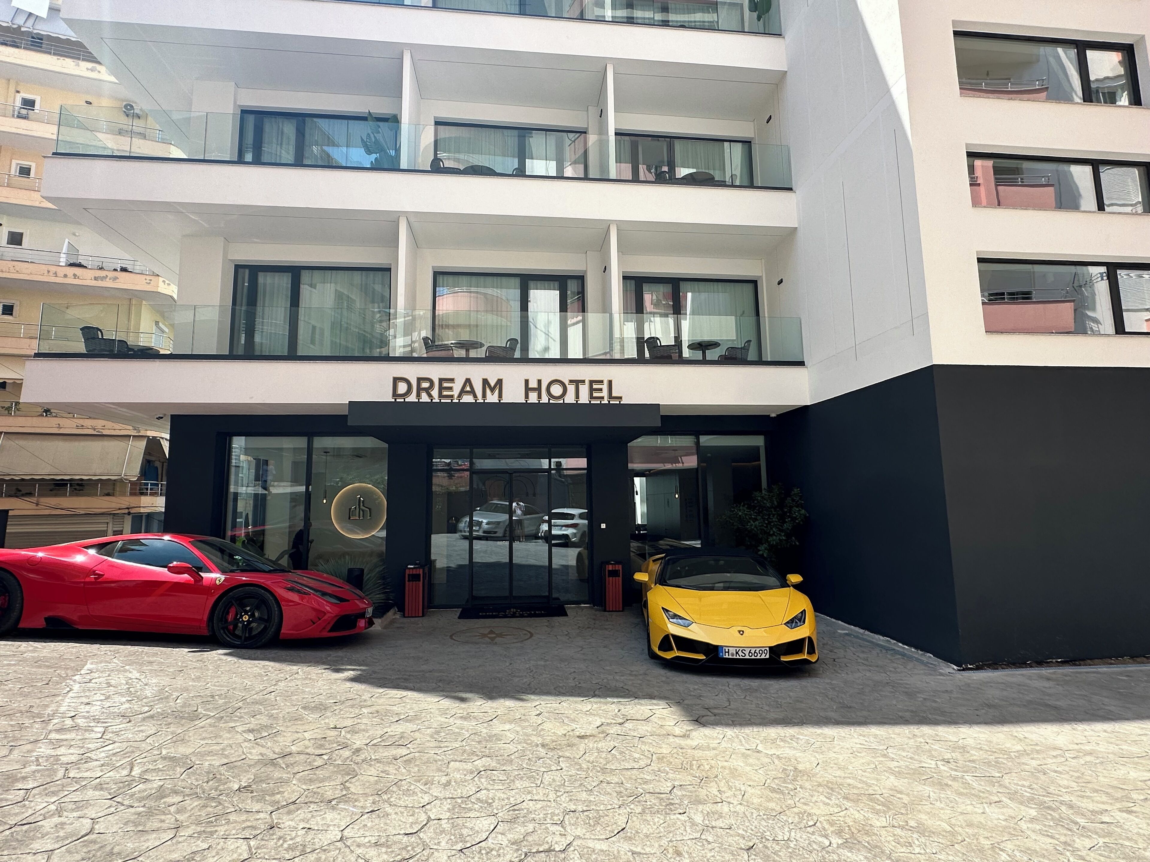 A modern hotel with a prominent "DREAM HOTEL" sign, featuring a glass facade, a red sports car, and a yellow sports car parked in front.
