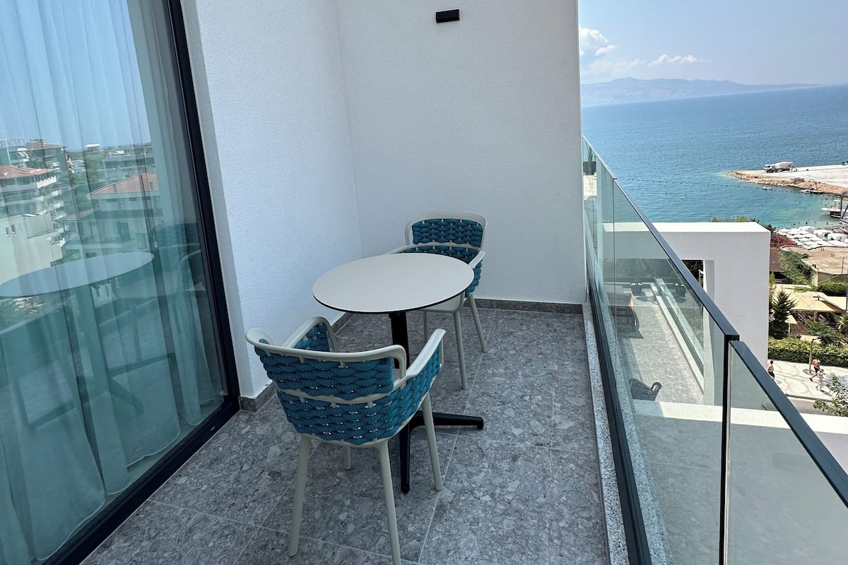 Ein Balkon mit Glastisch und Stühlen, freier Blick auf das Meer und eine weiße Wand.