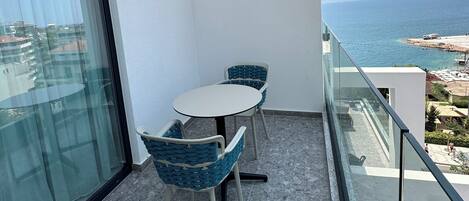 A balcony with a glass table and chairs, a clear view of the sea, and a white wall.