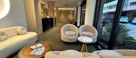 A modern hotel lobby with a reception desk, seating area with sofas and armchairs, and a large window offering a view of the street.