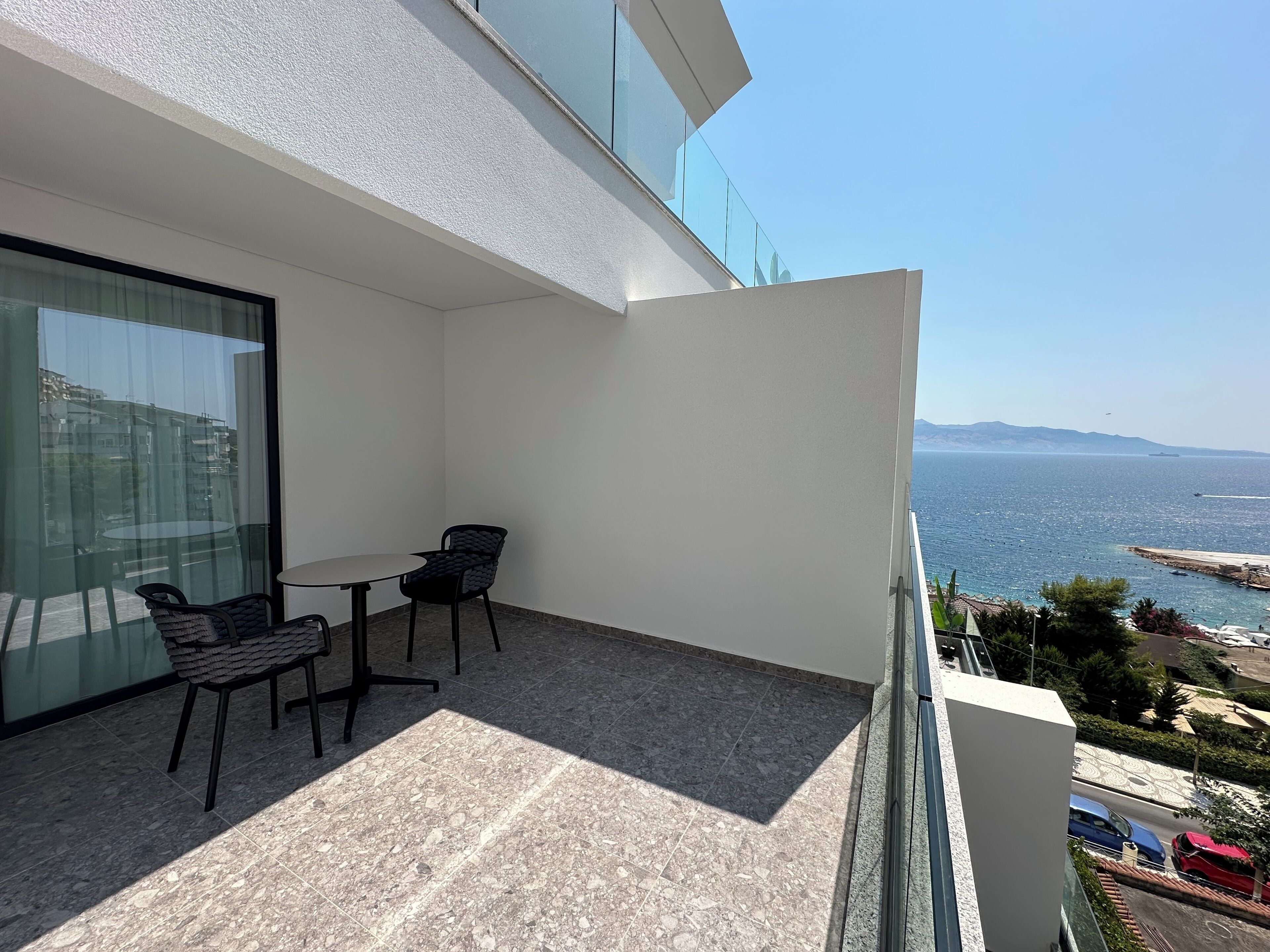 A balcony with a table and chairs, a glass door, and a view of the sea and distant mountains.