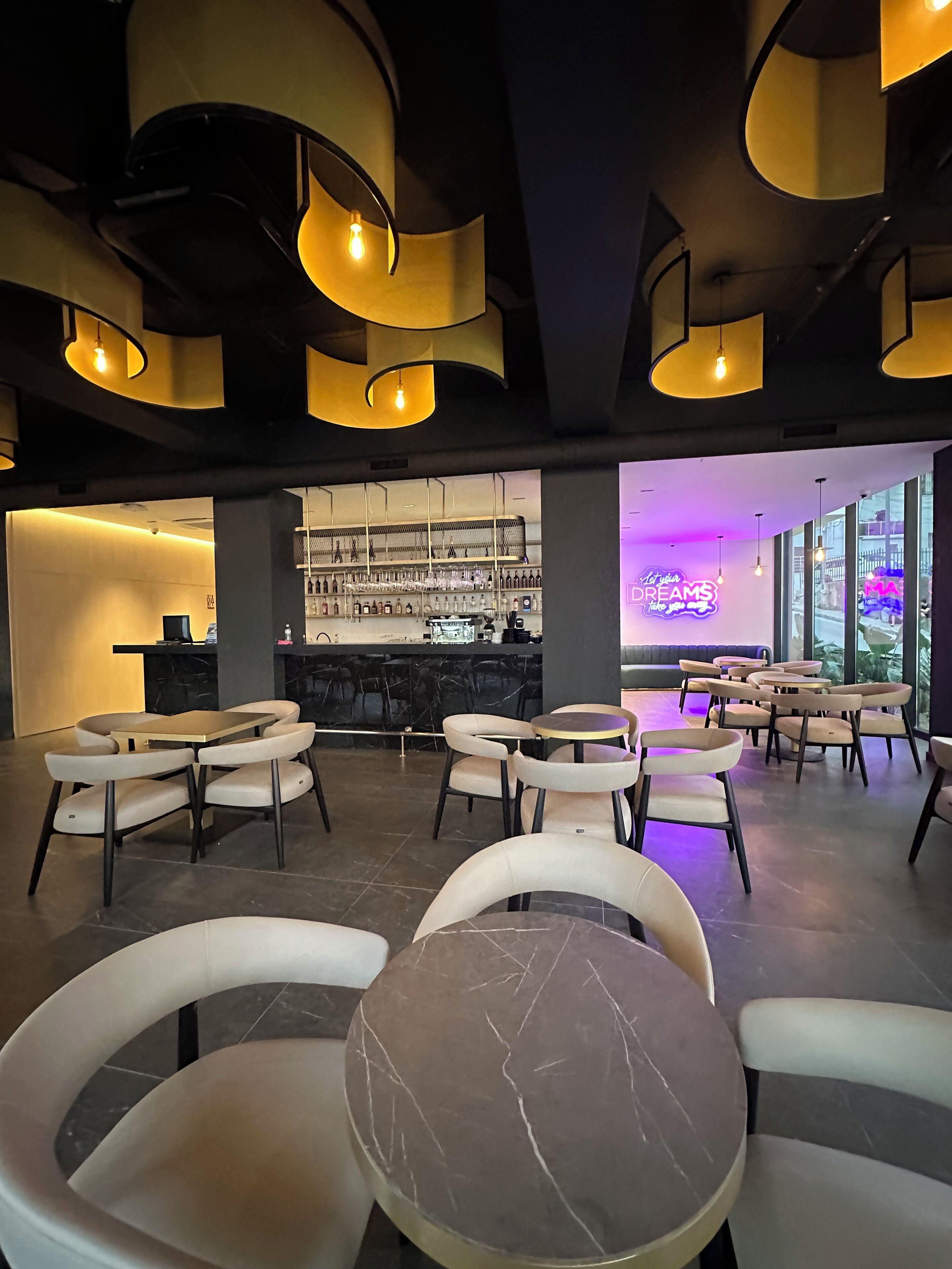 A modern bar area with marble-topped tables and chairs, a bar counter with a bartender, and a neon sign in the background.