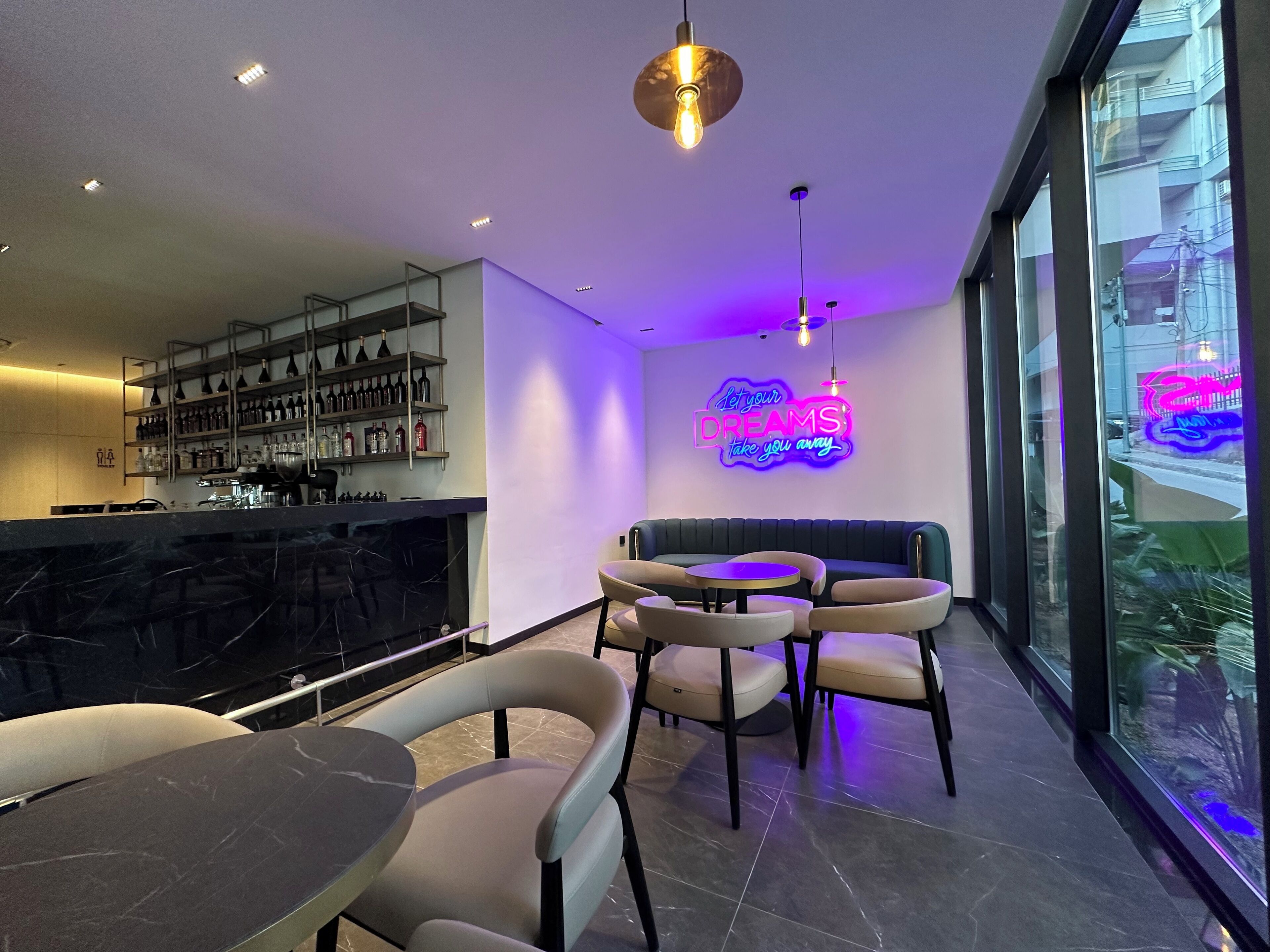 A modern bar area with a neon sign, seating, and a view of the city through large windows.