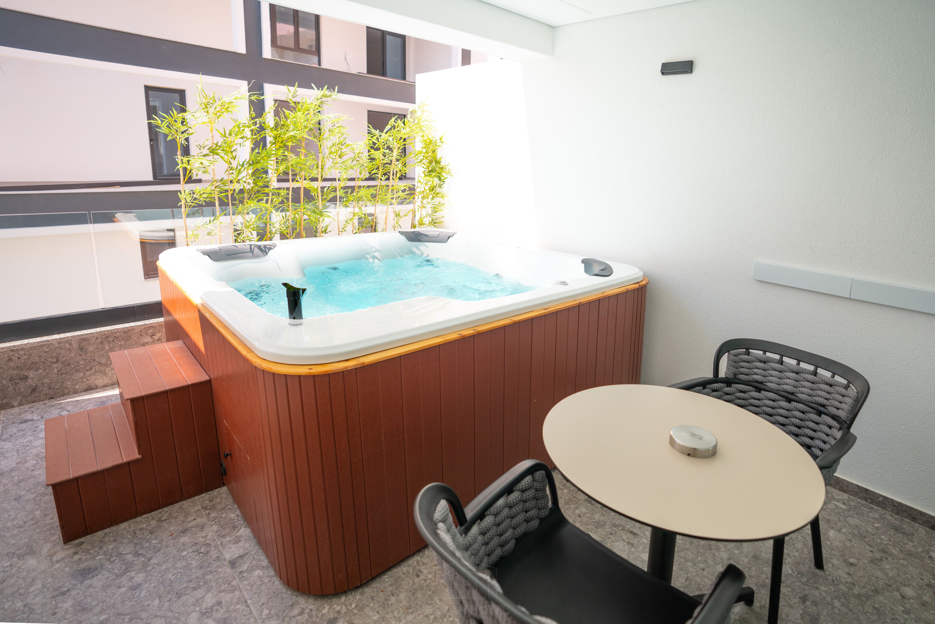 Un jacuzzi con revestimiento de madera, una mesita pequeña y dos sillas en un balcón con vistas a edificios y árboles.