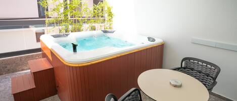 A hot tub with a wooden exterior, a small table, and two chairs on a balcony with a view of buildings and trees.
