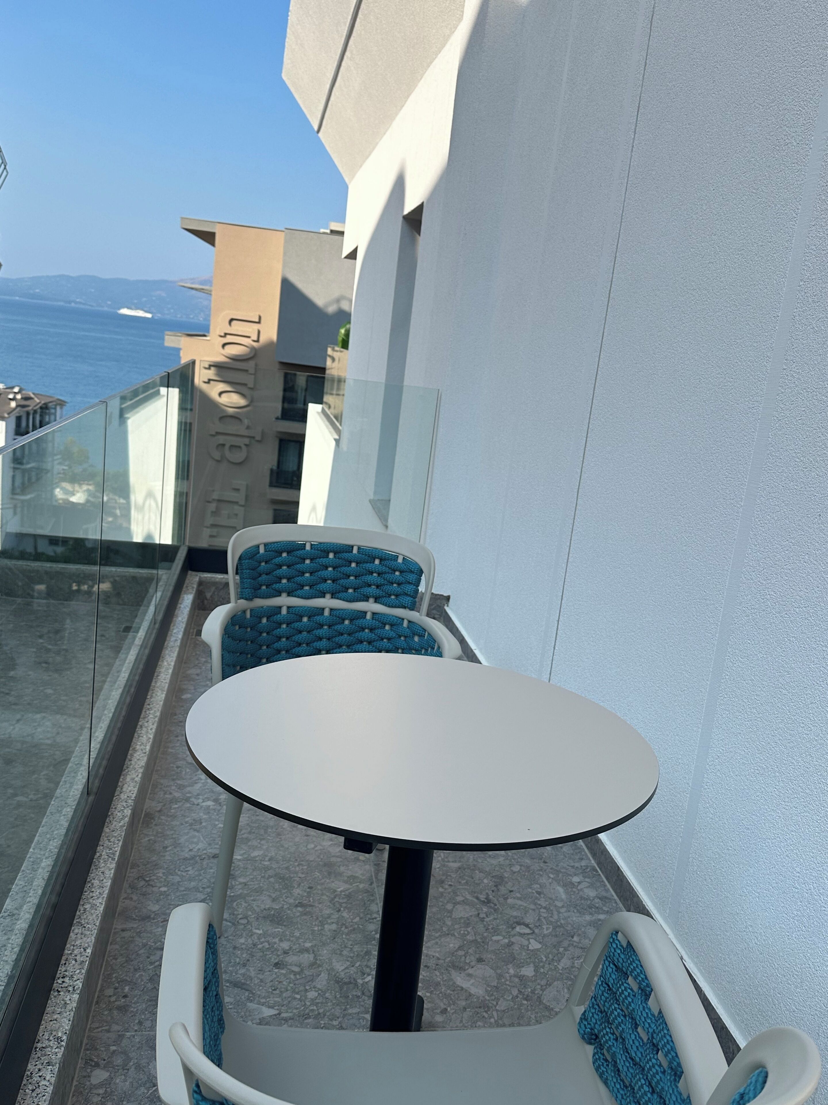 Un balcon avec une table ronde et deux chaises bleues, donnant sur une étendue d’eau et des bâtiments.
