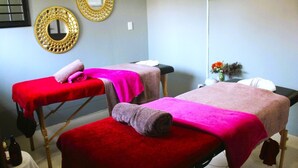 A room with two massage tables, each covered with a red towel and a pink blanket. There are decorative mirrors on the wall and a small table with a vase of flowers.