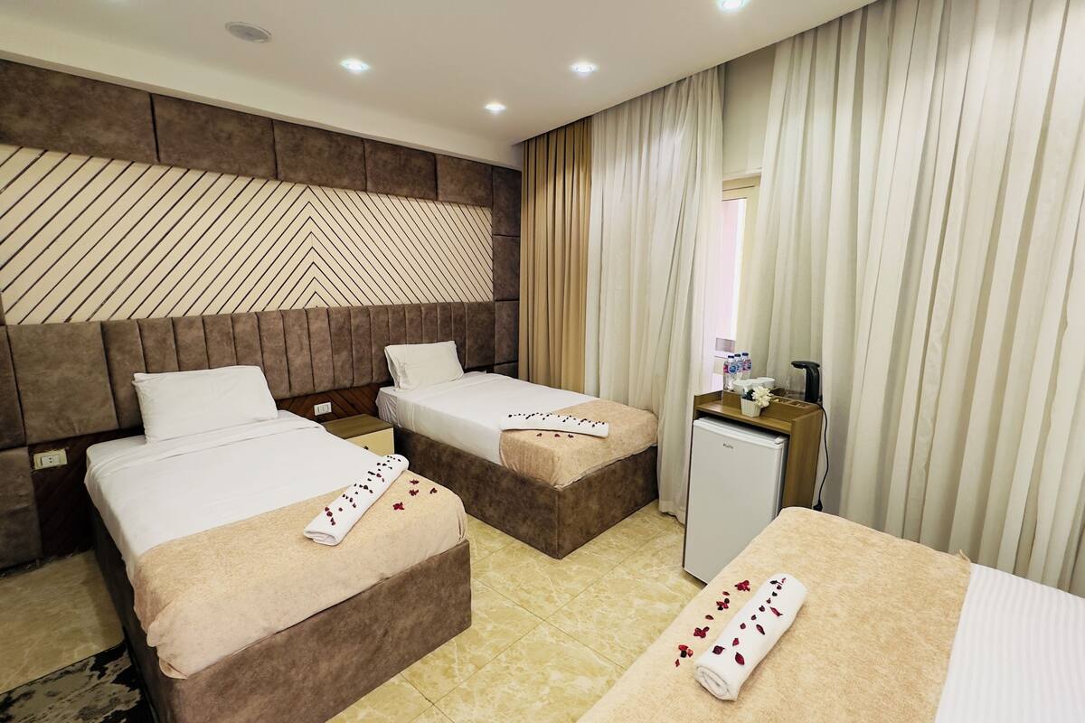 A hotel room with two beds, a minibar, and a large window with curtains.