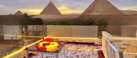 An outdoor dining area with a table set for two, surrounded by rose petals and a 'LOVE' sign. In the background, there are two pyramids and a sunset sky.
