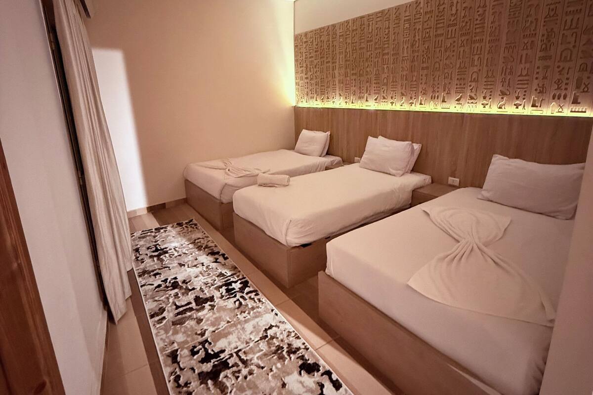 A hotel room with a modern design, featuring two single beds and a large bed, a patterned carpet, and a decorative wall panel.