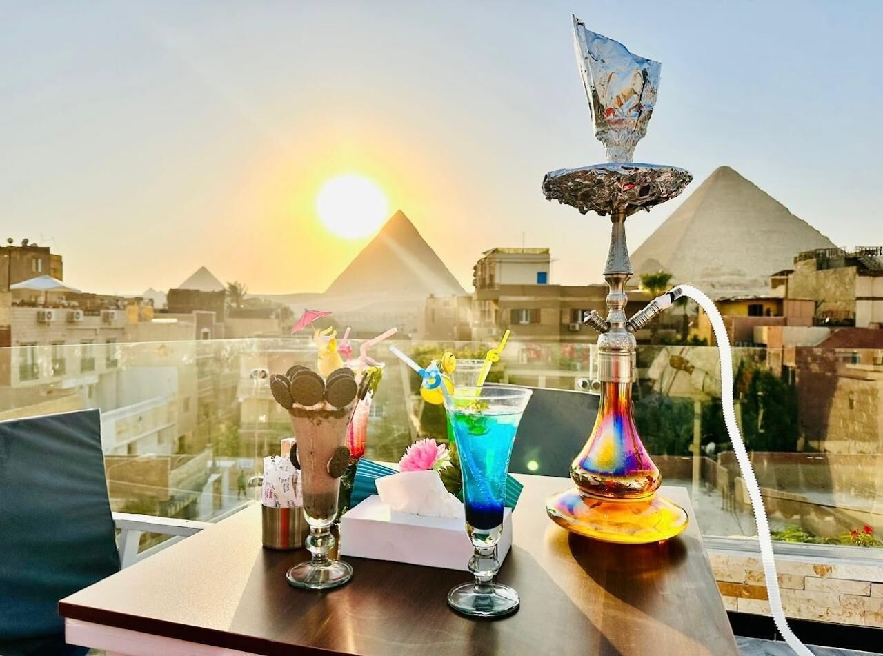 A table with drinks and a hookah, overlooking a cityscape with pyramids in the background.