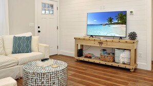 A living room with a white sofa, a round coffee table, a wooden TV stand with a TV displaying a beach scene, and a door with a window. - The Surf Ranch: Near Bay, Pet Friendly! (Port St. Joe)