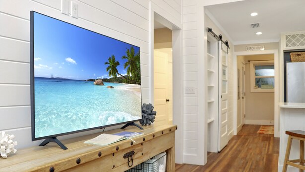 A TV on a wooden console with a beach scene, a white interior, and a 'Beach House' sign. - The Surf Ranch: Near Bay, Pet Friendly! (Port St. Joe)