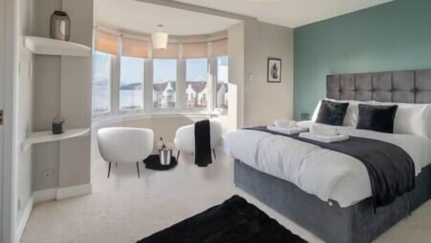 A modern bedroom with a large bed, a view of the city, and a small seating area. - Penthouse at the Bay near Edinburgh (Dalgety Bay)