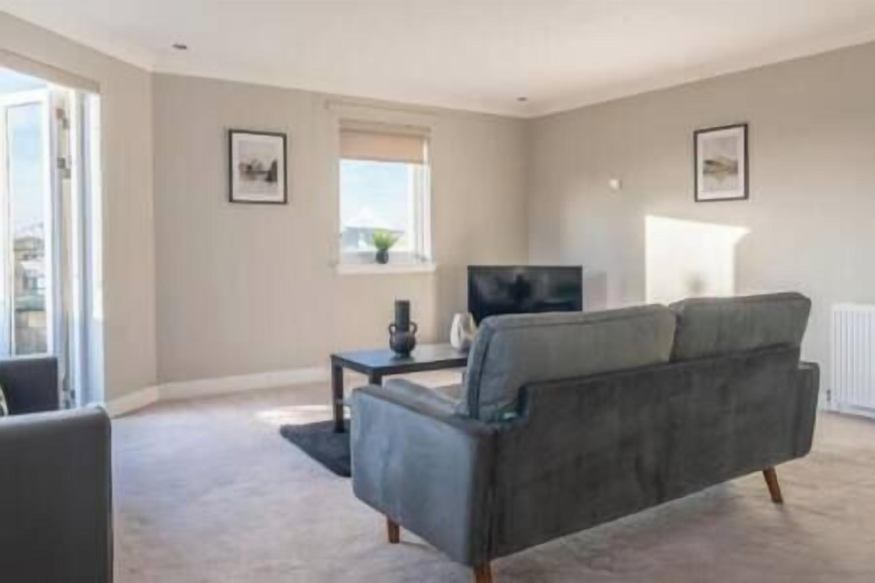 A living room with a grey sofa, a small table, a TV, and a window with blinds.