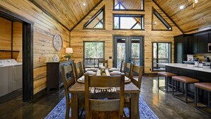 A wooden cabin interior with a dining area, kitchen, and a large window overlooking the outdoors.