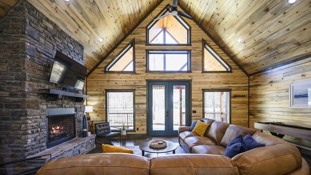 A cozy log cabin interior with a stone fireplace, leather sofa, and a high ceiling with wooden beams.