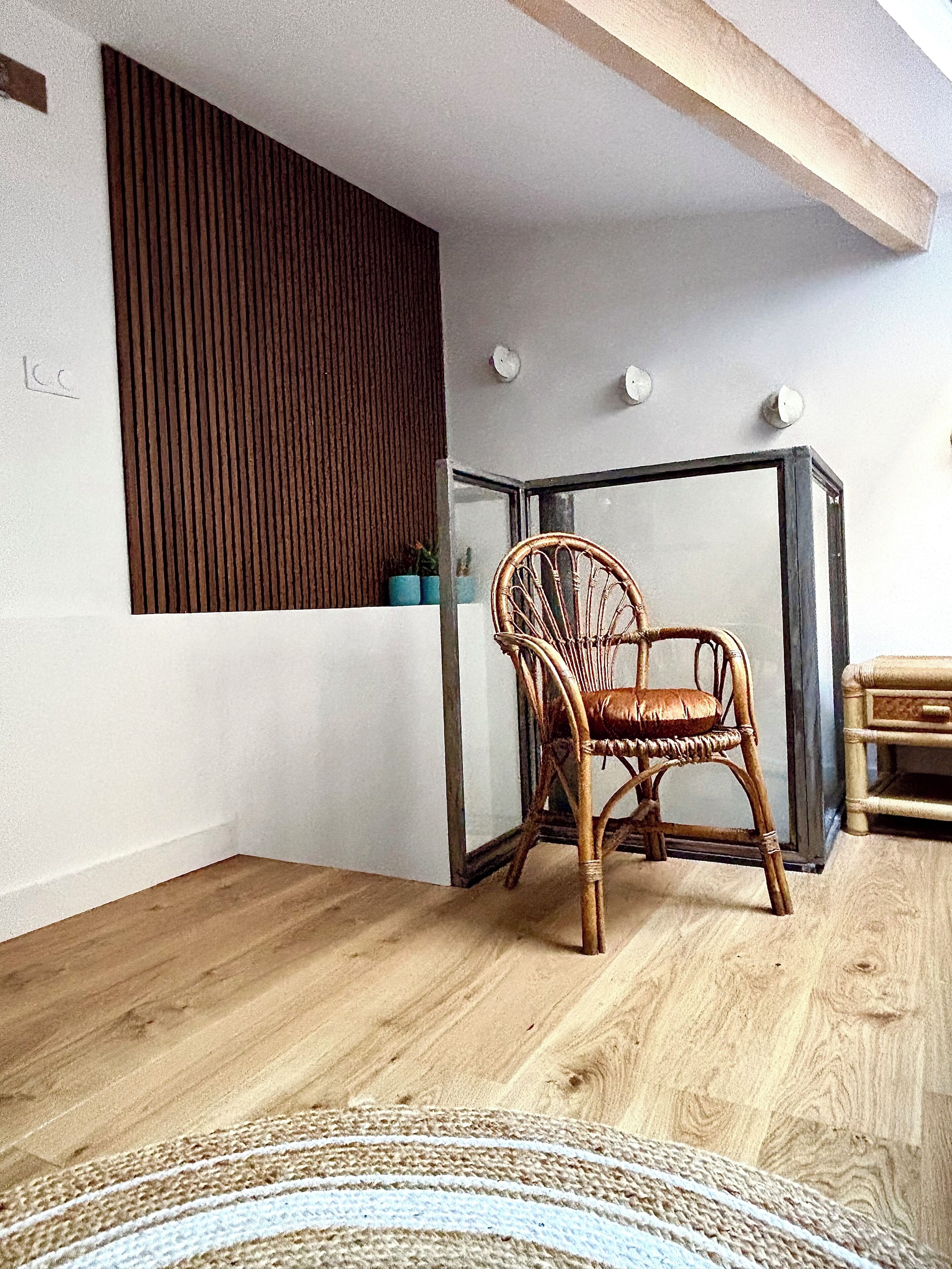 A room with a wooden floor, a wicker chair, a glass table, and a white wall with a wooden panel.
