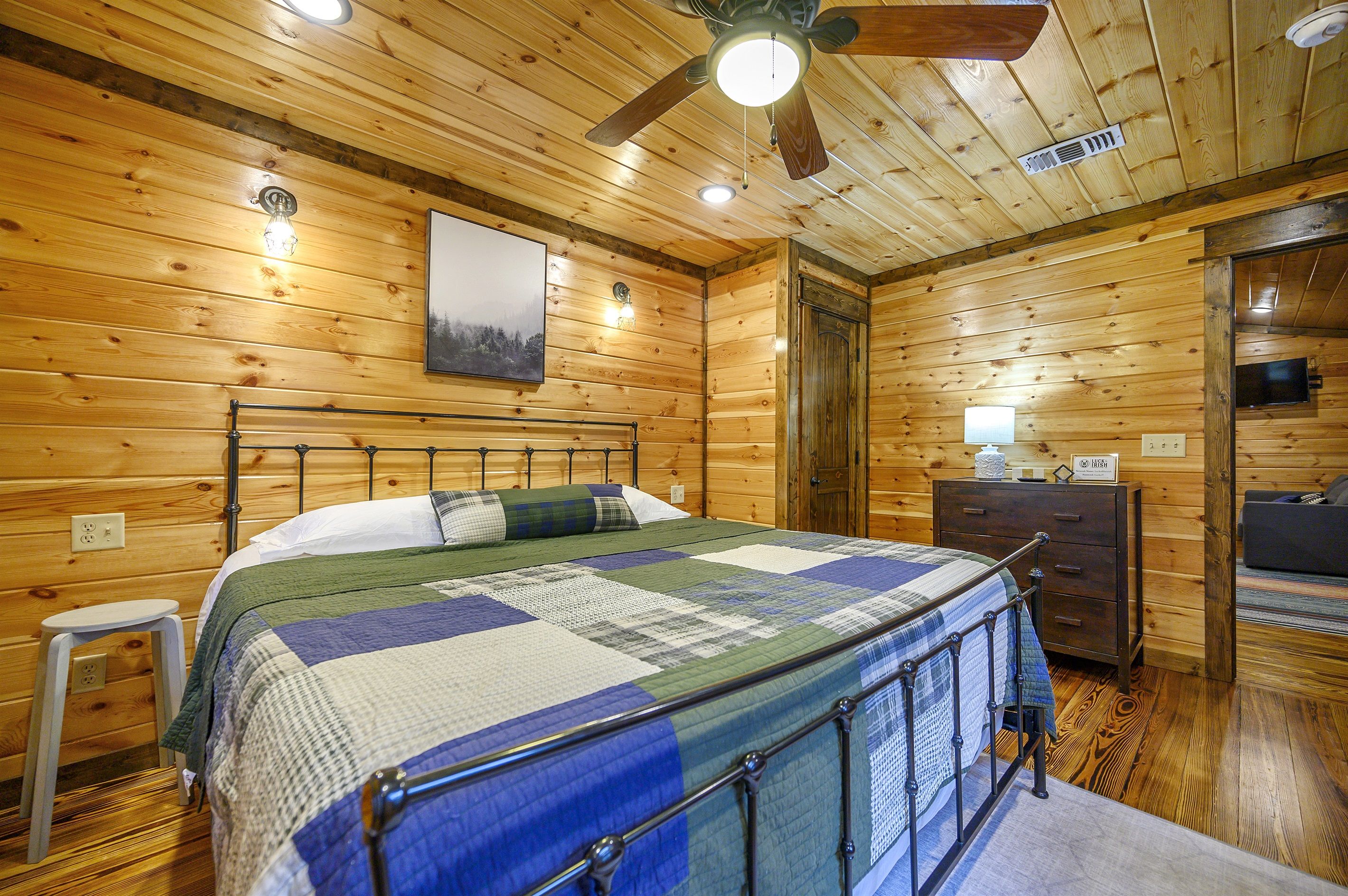 Une chambre à coucher aux murs lambrissés, comprenant un lit, une table de chevet, une commode et un ventilateur de plafond.
