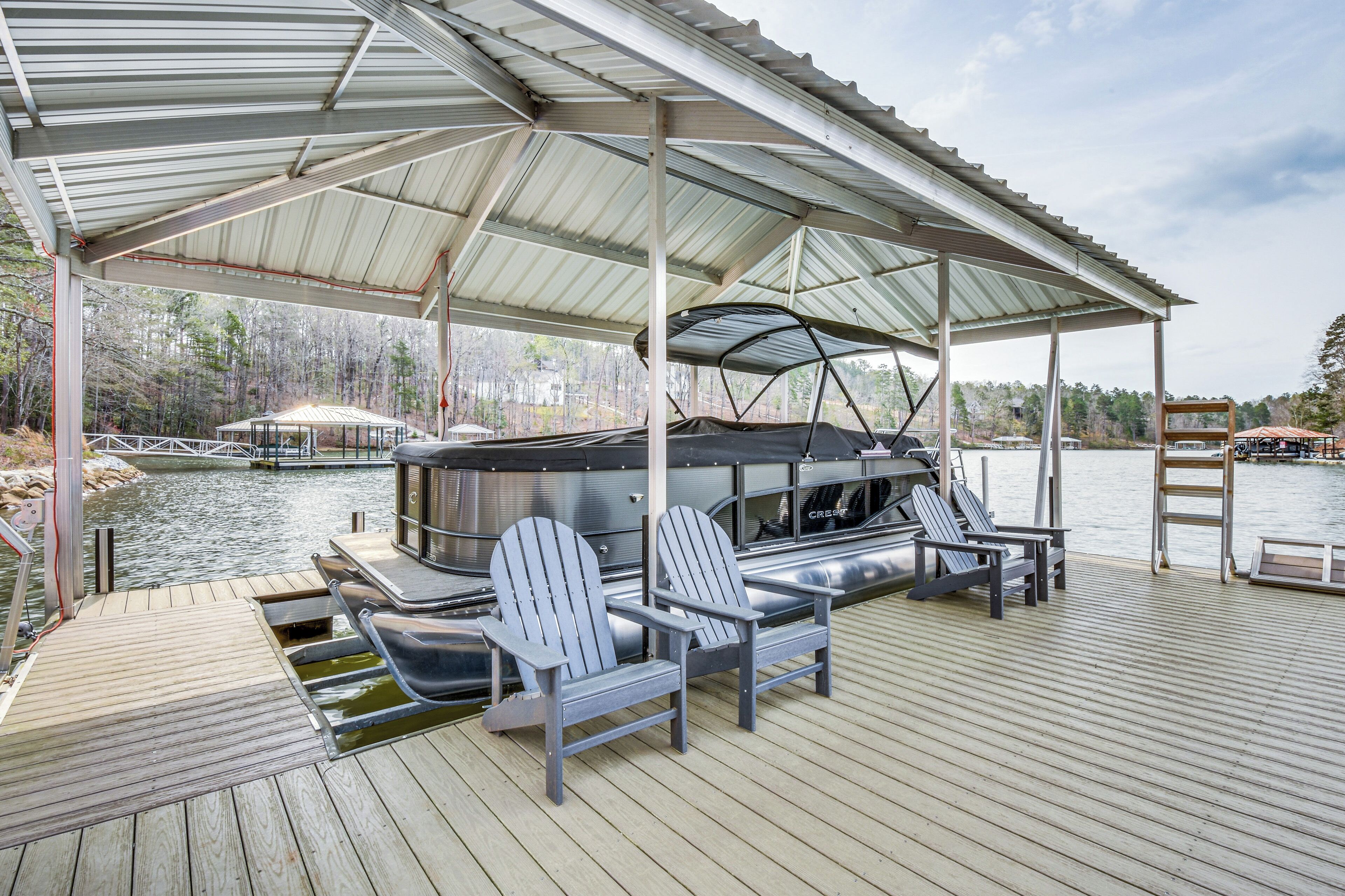 Um deck coberto com cadeiras Adirondack, uma churrasqueira e um barco atracado ao fundo.