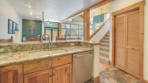A kitchen with granite countertops, wooden cabinets, a dishwasher, and a view of a staircase leading to a second floor.