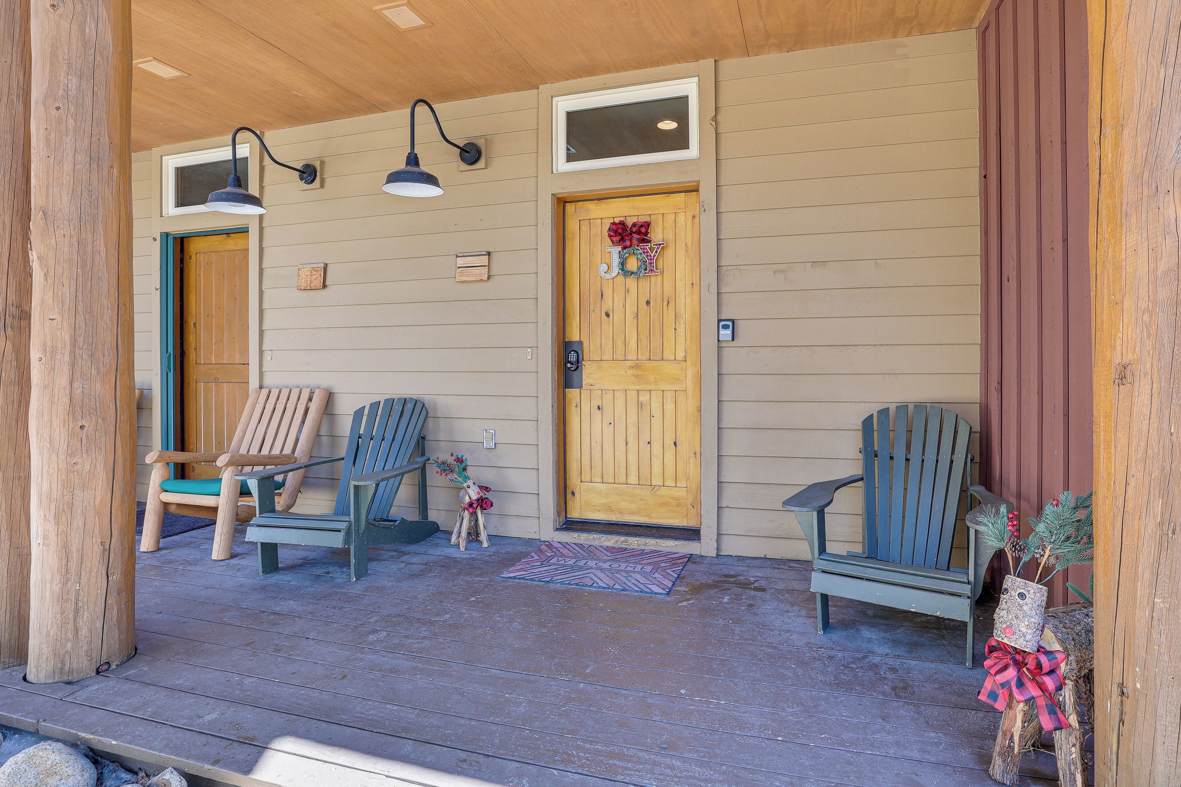A wooden porch with two Adirondack chairs, a wooden door with a 'JOY' sign, and a small wooden table.