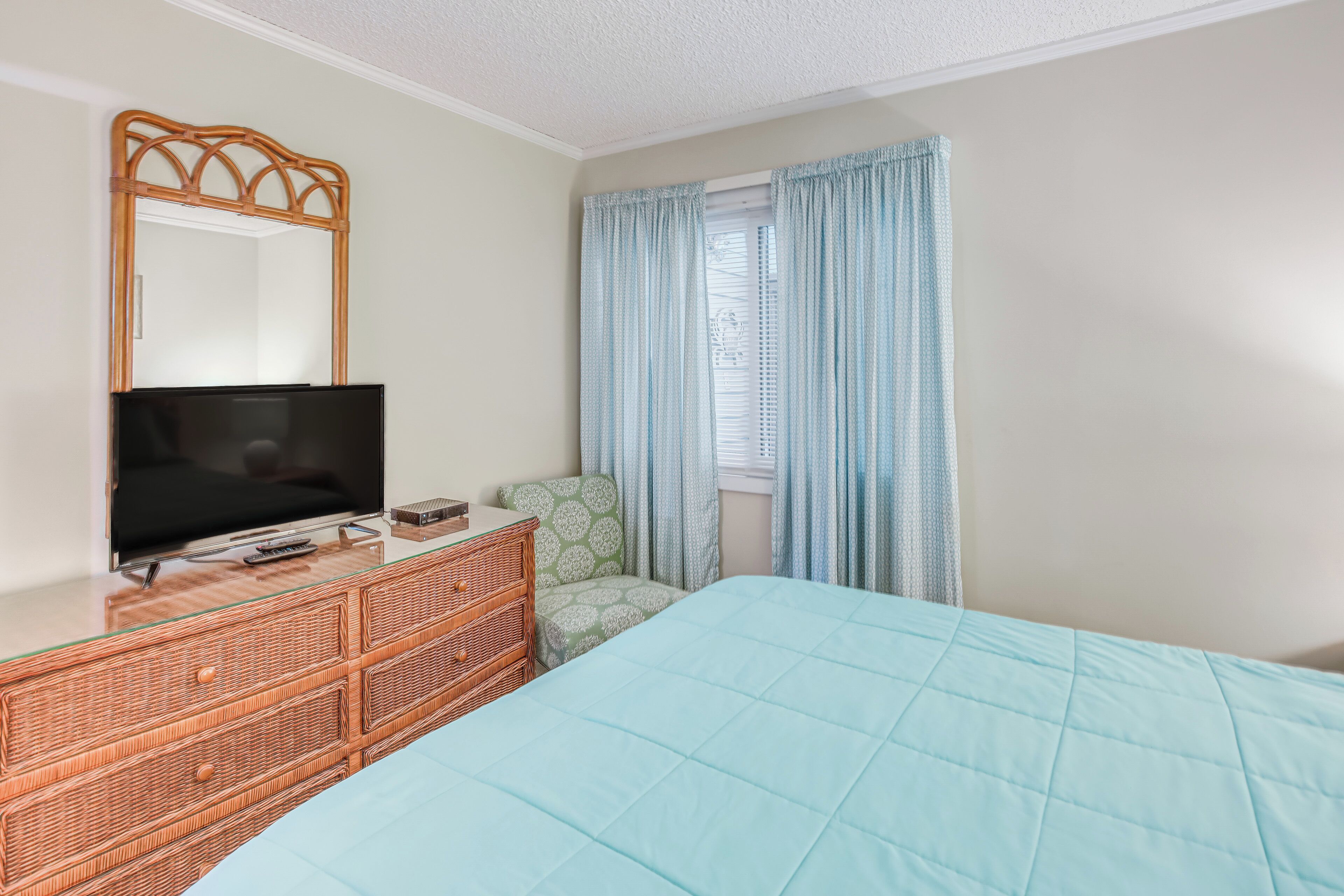 A bedroom with a television on a glass-top dresser, a bed with a blue quilt, and light blue curtains.