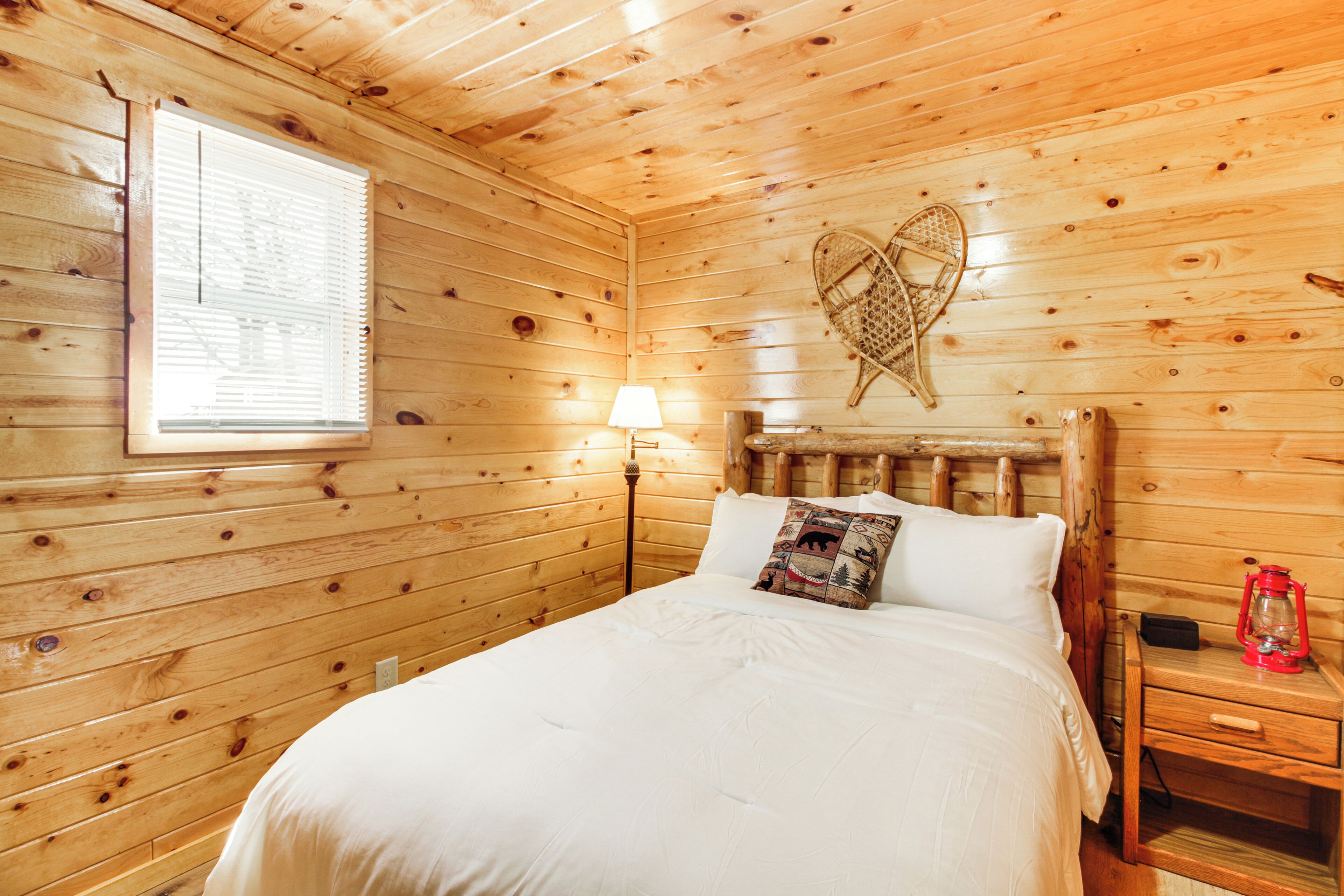 Una habitación de cabaña de madera con una cama, una mesita de noche, una lámpara y una ventana con persianas.